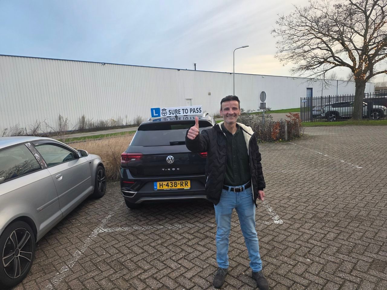 Geslaagde leerling met opgestoken duim voor de lesauto op het parkeerterrein