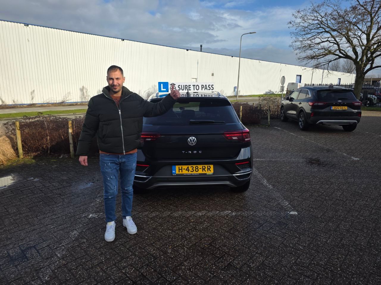 Geslaagde leerling staat naast de lesauto met het L-bord op de achterklep